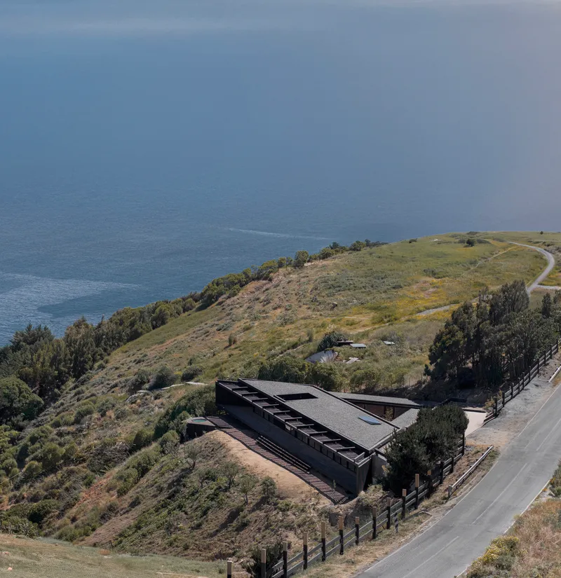 Aerial view of the angular roof form nestled into coastal hillside terrain with ocean in background