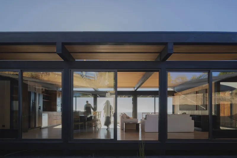 Exterior view through glazed facade showing interior spaces with timber ceiling and figures moving at dusk