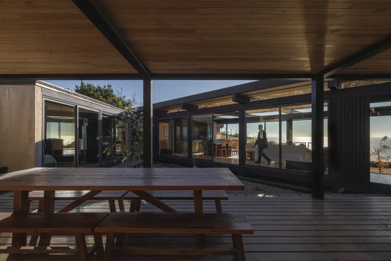 Covered timber deck with picnic table looking through glazed volumes toward the sea