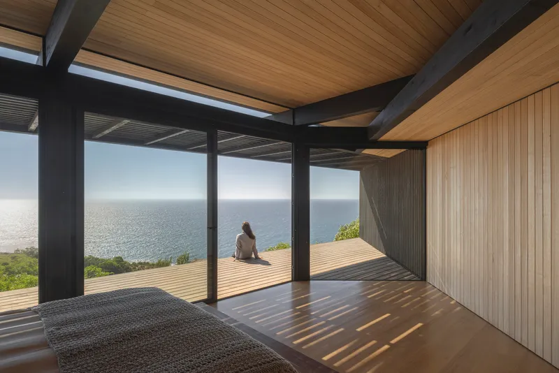 Interior room with timber-clad walls and ceiling looking out to a wooden deck overlooking the ocean with a seated figure