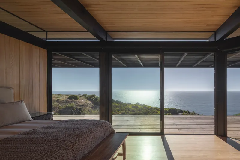 Bedroom with exposed steel beams and timber ceiling opening to glazed walls facing the coastal landscape at midday