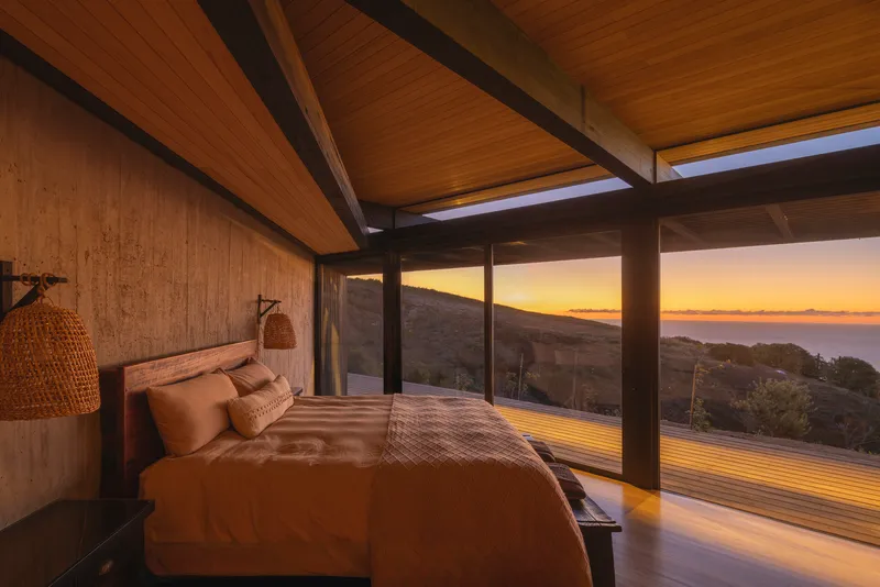 Bedroom with timber walls and ceiling framing floor-to-ceiling windows overlooking the coastline at sunset