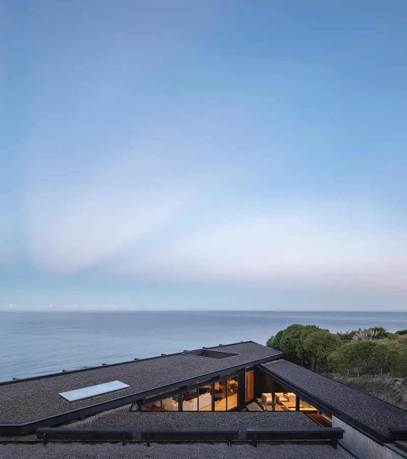 Angled gravel roof with skylights and glazed openings set into coastal vegetation at dusk