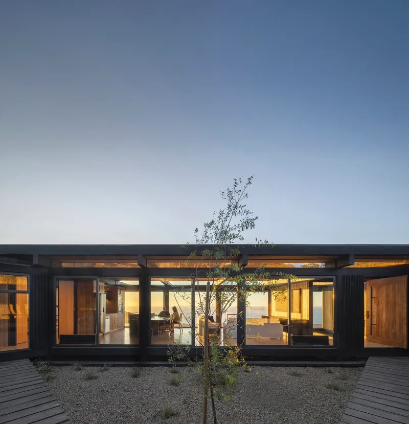 Glazed courtyard facade with steel frame and central tree surrounded by gravel at twilight