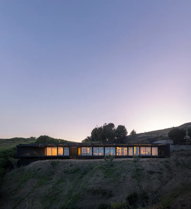 Illuminated linear volume set into a hillside viewed at dusk with rolling terrain and trees beyond