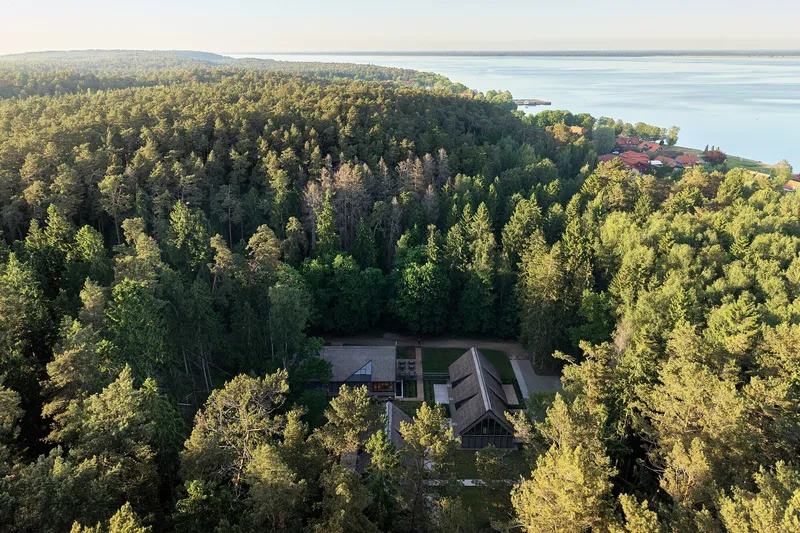 Aerial view of three gabled volumes nestled in a forest clearing near a lakeshore
