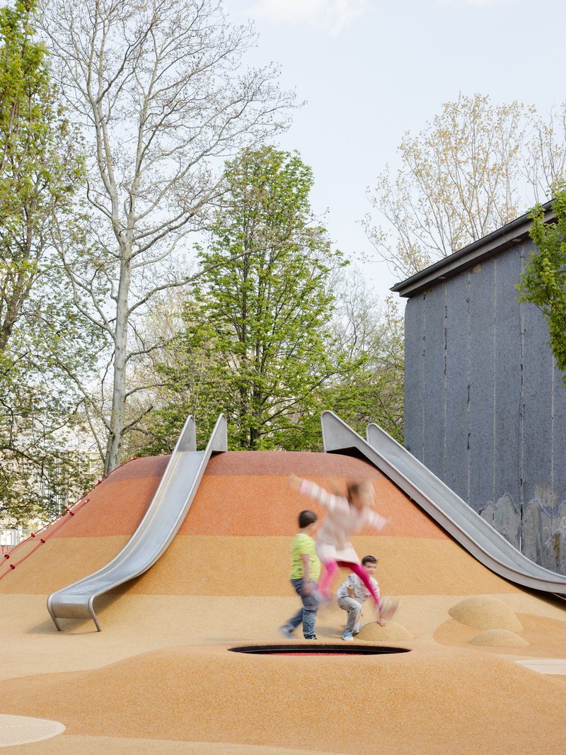 Mounded orange play surface with stainless steel slides and children running near concrete wall