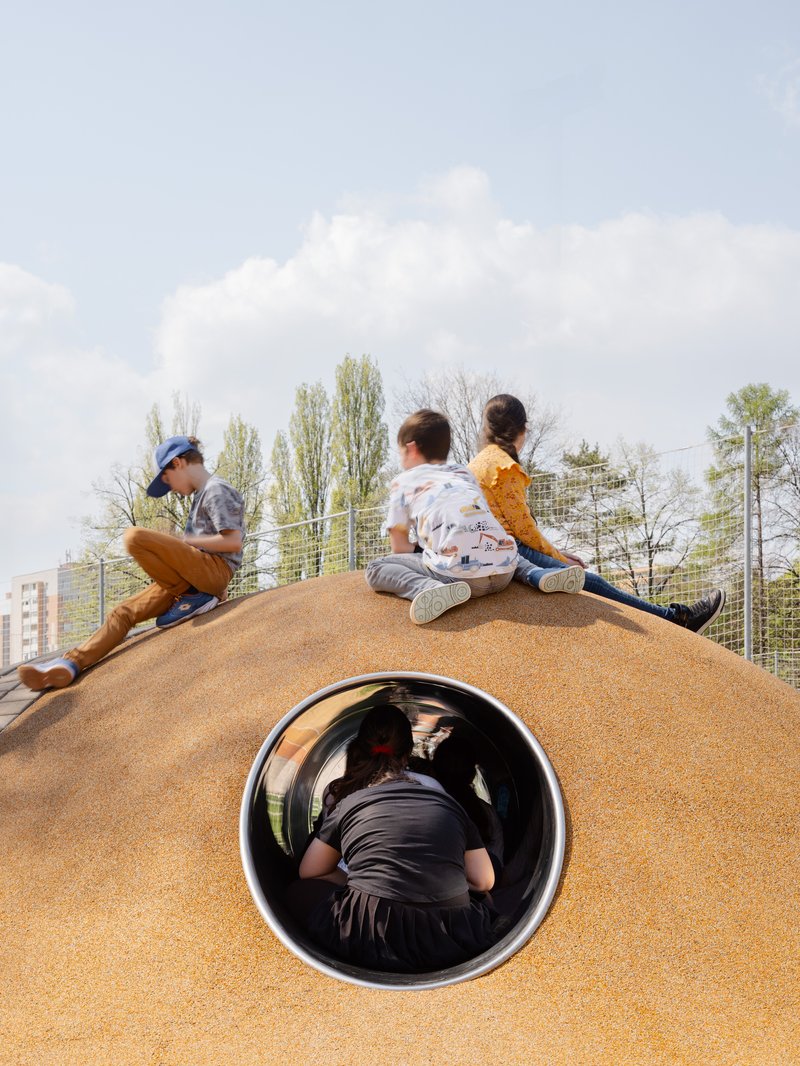 Children sitting on orange mound above black tubular tunnel opening under cloudy sky