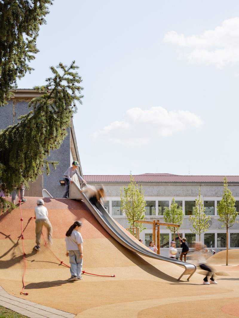 Sloped orange play surface with metal slide and children under evergreen branch