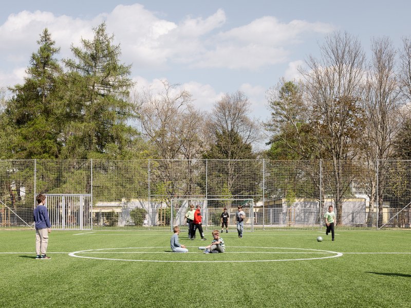 Artificial turf football pitch with metal mesh fencing and children playing under a partly cloudy sky