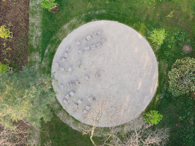 Overhead view of a circular concrete amphitheater with timber seating benches among deciduous trees