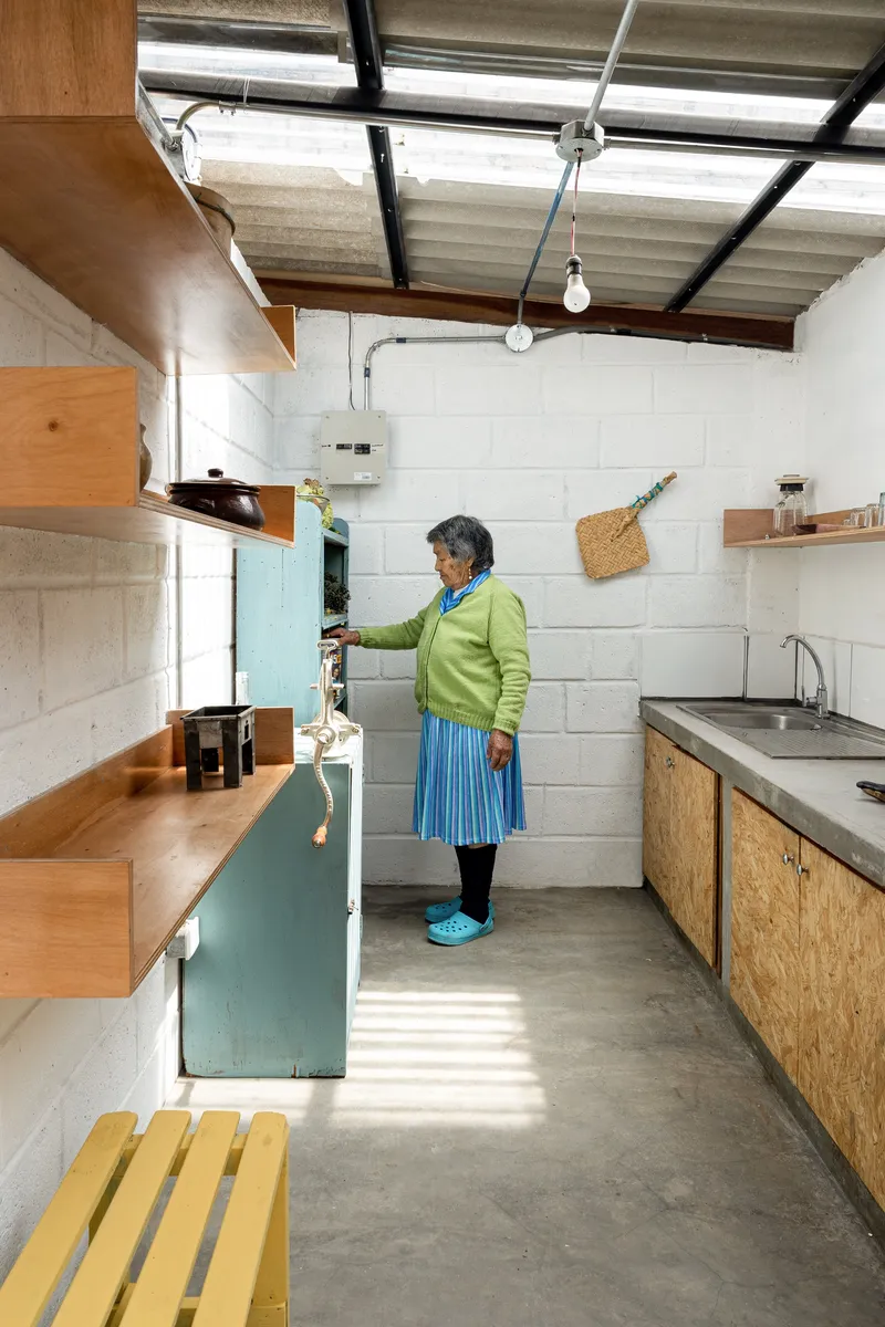 Compact kitchen with OSB cabinetry and a woman standing at the refrigerator under natural light