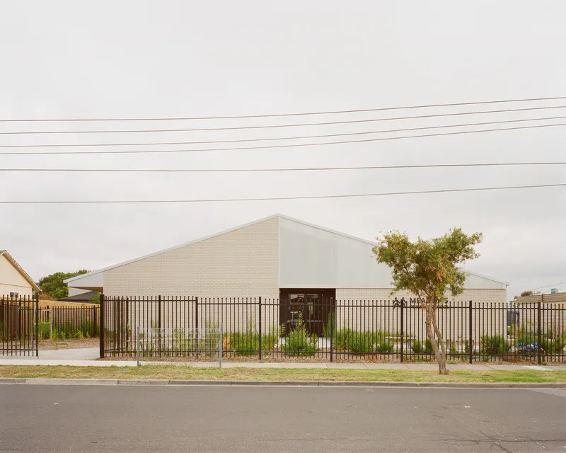 Street view of the white brick gabled volume behind vertical steel fencing and young trees