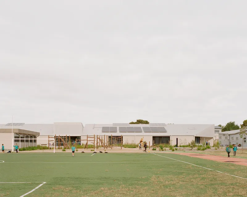 View across sports field showing low-slung building with solar panels on sawtooth roofs and children playing on grass
