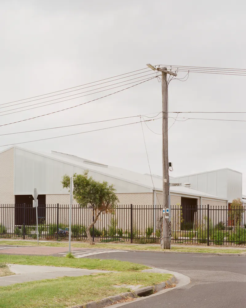 Street view showing pale brick and metal roofed volumes behind security fence with utility pole in foreground