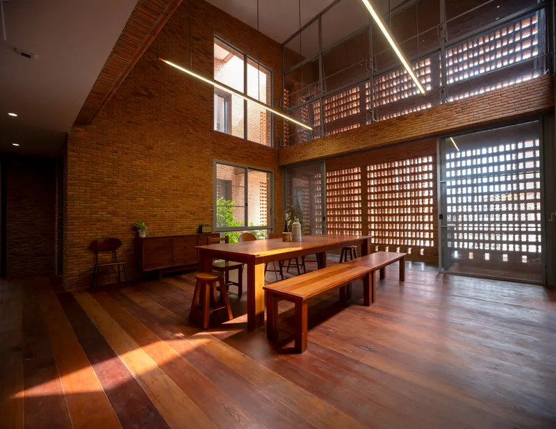 Double-height dining space with timber table, benches, and perforated brick screens filtering daylight