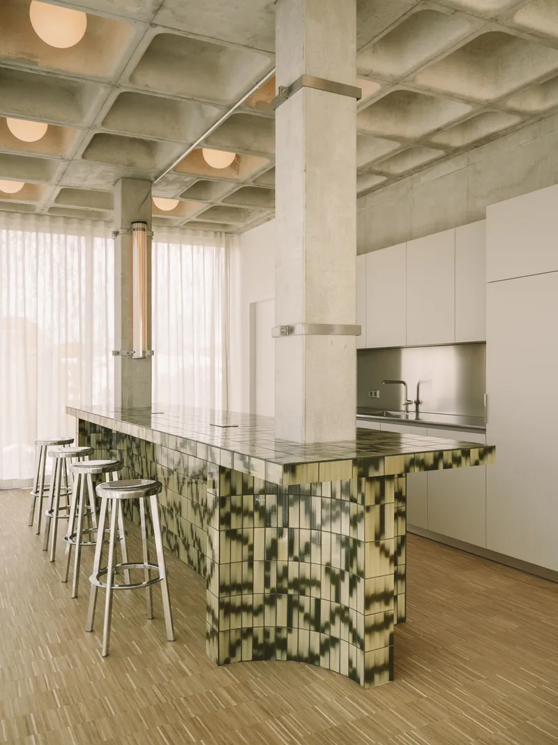 Kitchen island clad in variegated green tile beneath waffle concrete ceiling with spherical pendant lights