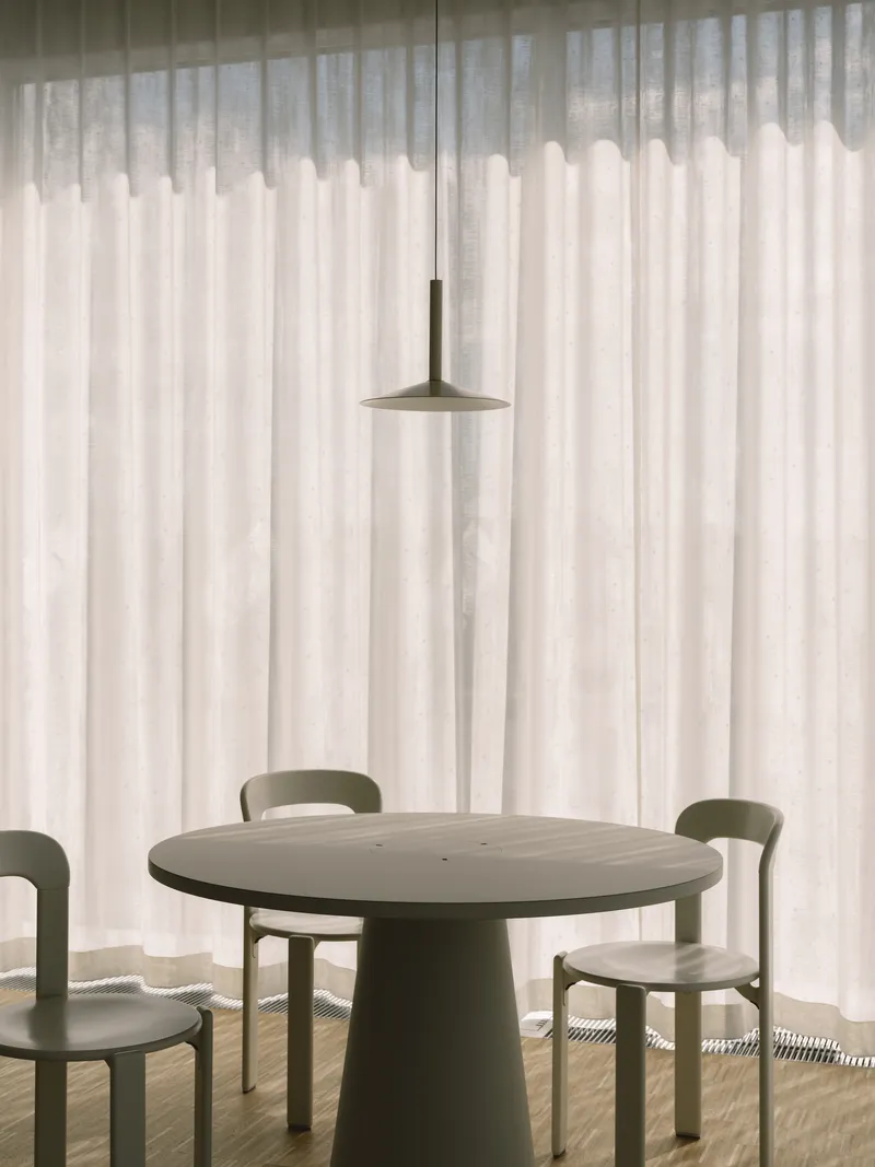 Round dining table with pedestal base and thin pendant light against floor-to-ceiling sheer curtains in soft daylight
