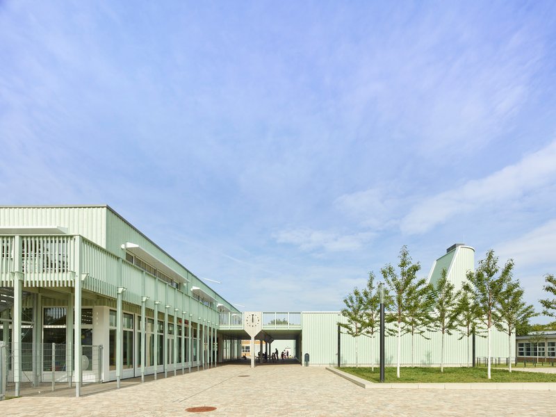 Paved courtyard flanked by pale green corrugated metal facades with white columns and young trees
