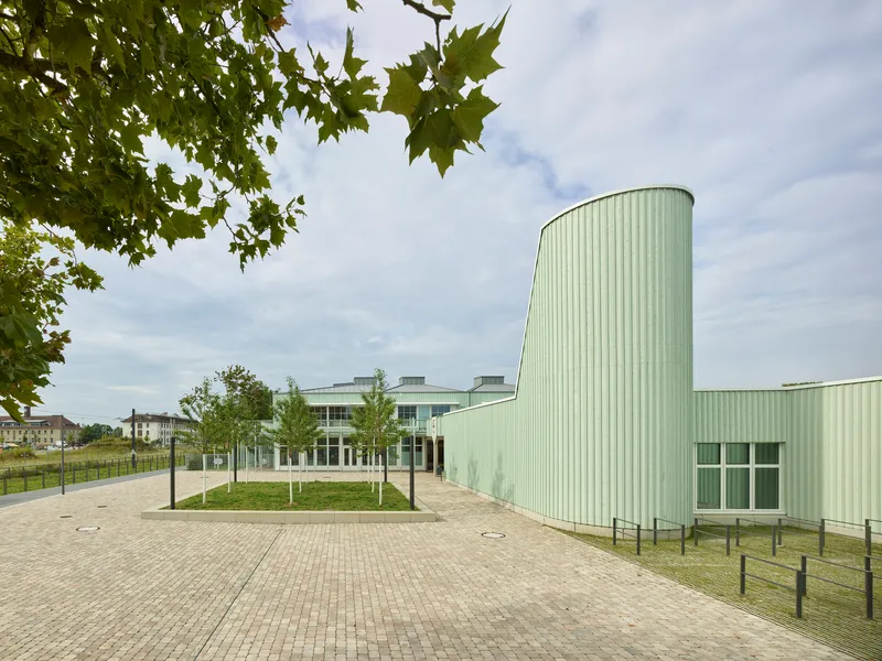 Curved pale green corrugated metal tower beside a plaza with young trees framed by foliage