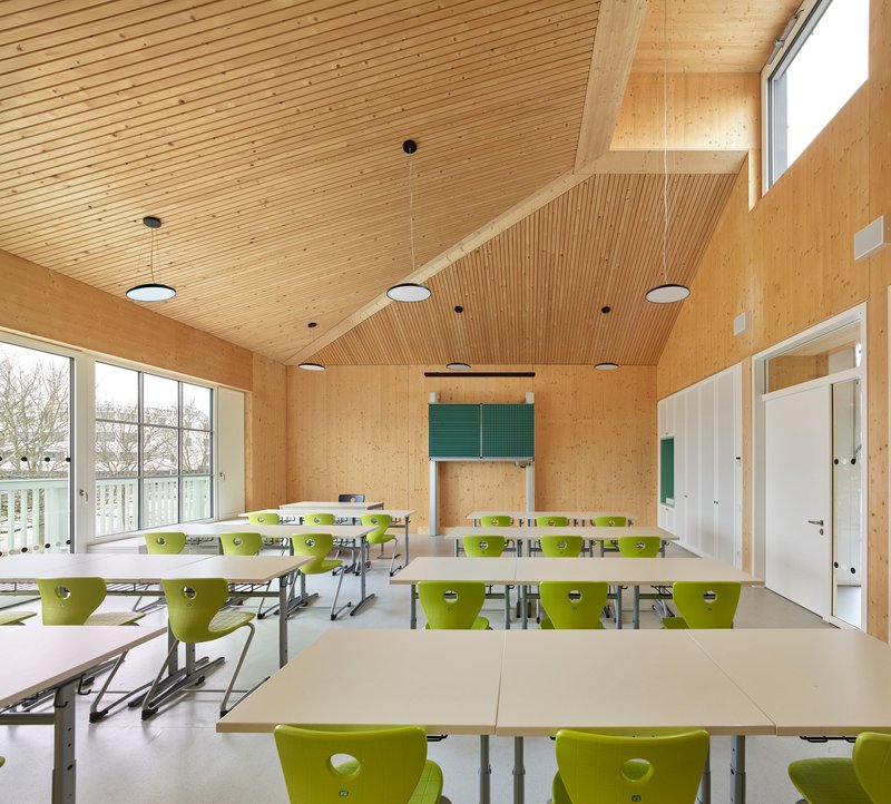 Classroom interior with green chairs and plywood walls beneath an angled timber ceiling