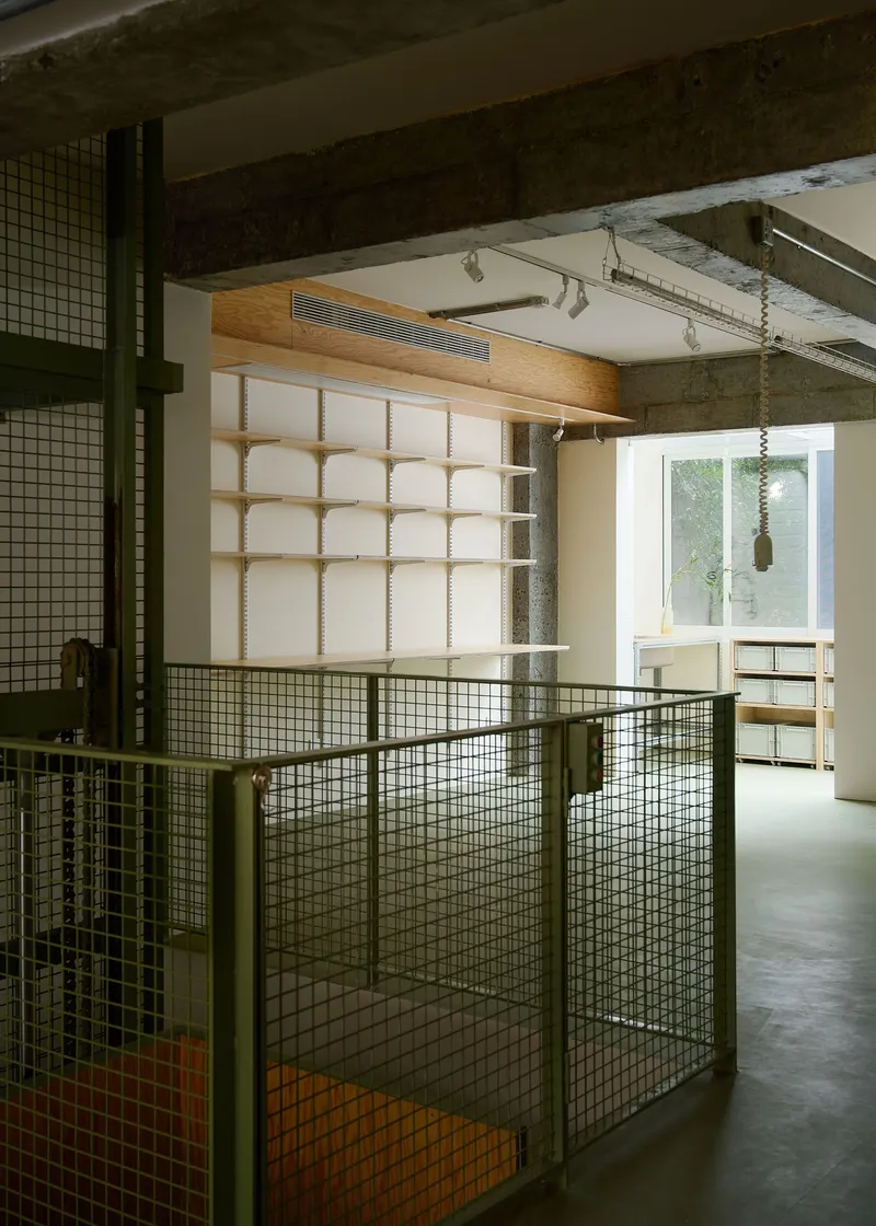 Interior view through green wire mesh partitions toward timber-framed shelving beneath exposed concrete beams