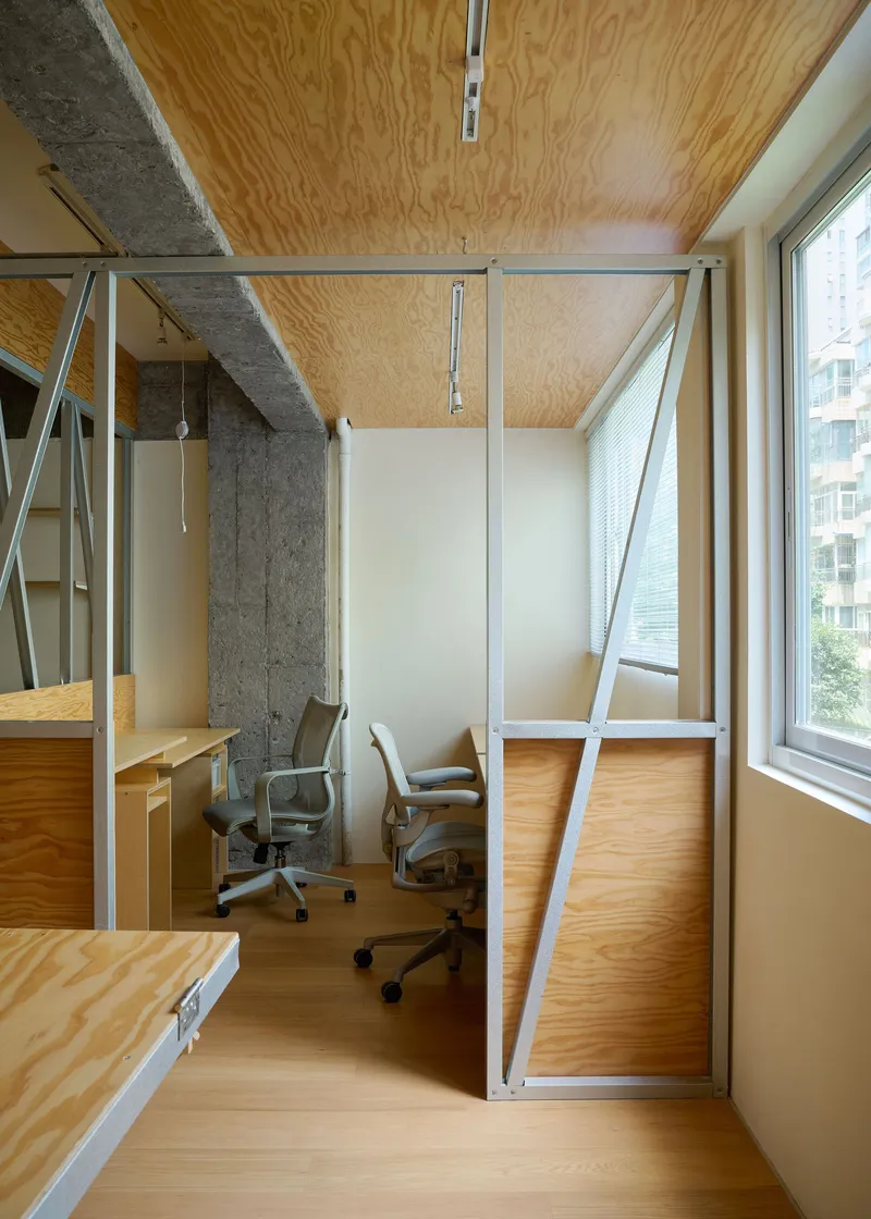 Enclosed workspace cubicle with steel framing and plywood paneling beneath a plywood ceiling and track lighting