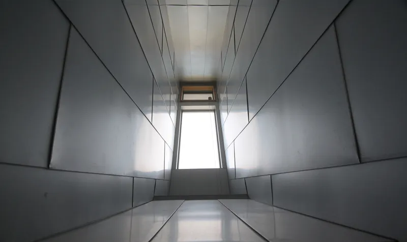 Narrow corridor lined with metal panels converging toward a distant window at the end