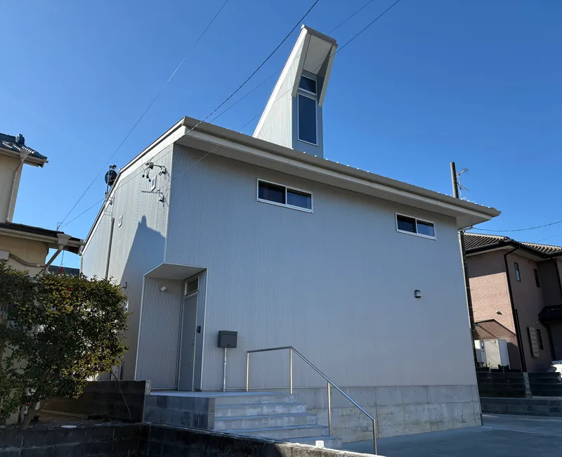 Corrugated metal exterior with vertical tower element and concrete entrance stairs among neighboring houses