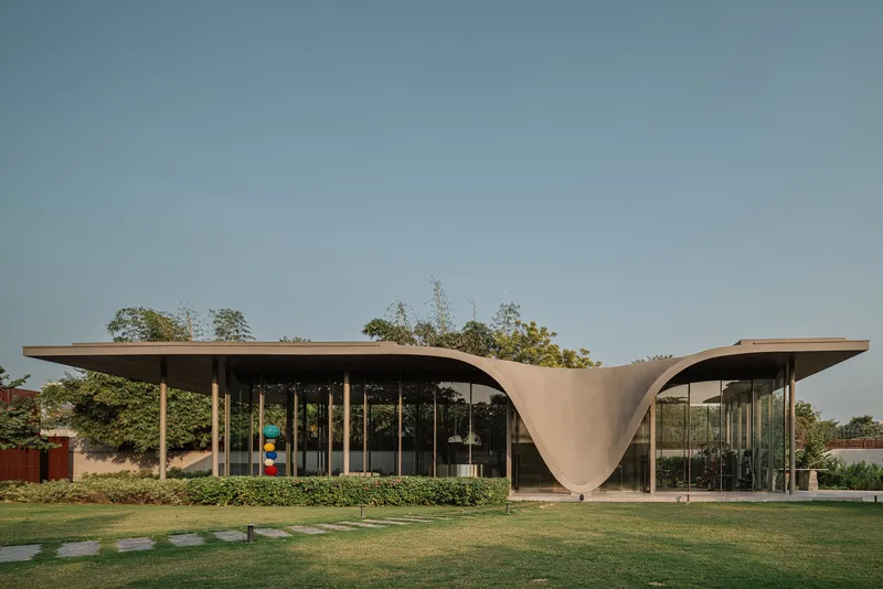 Glass pavilion with flowing concrete roof canopy set in a lawn with hedges and trees