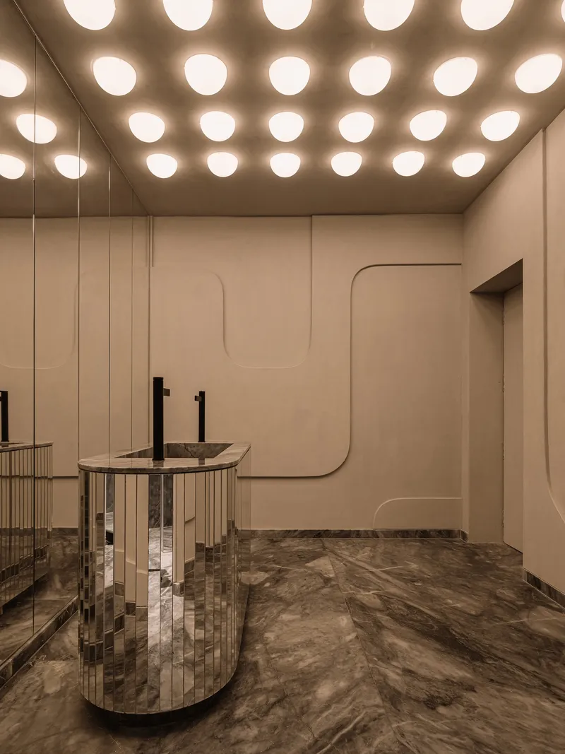 Bathroom with circular recessed ceiling lights above cylindrical mirrored sinks and sculpted wall panels in warm stone