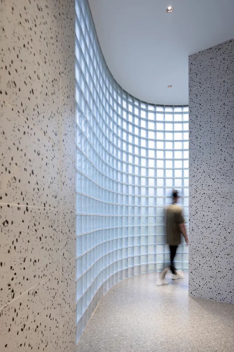 Curved glass block wall along a corridor with terrazzo panels and a visitor in motion