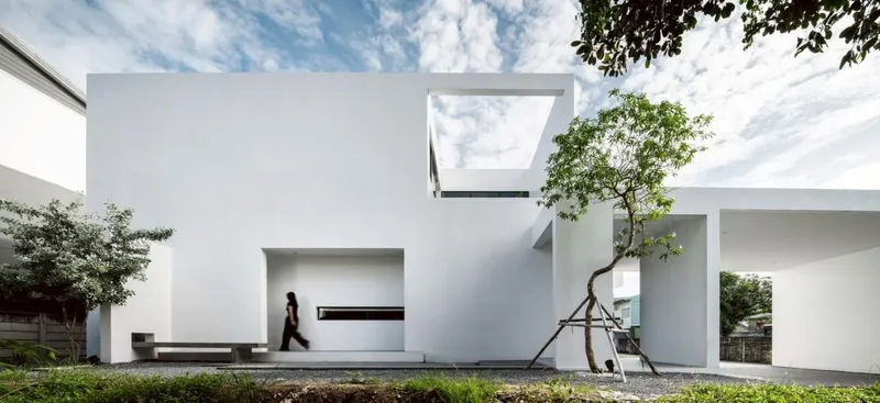 White cubic volumes with framed openings and a planted tree beside a gravel courtyard