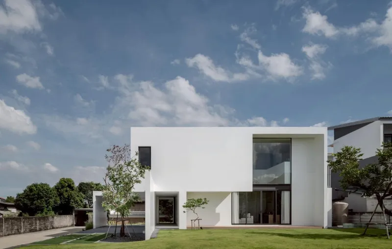 Rear elevation showing interlocking white volumes with glazed openings overlooking a lawn under scattered clouds