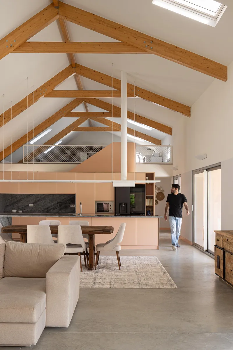 Double-height living space with exposed timber trusses, mezzanine, and person walking through kitchen area