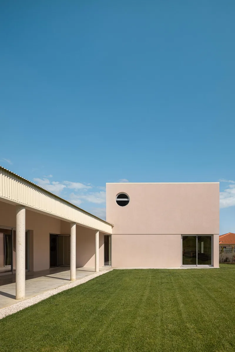 Garden facade showing pink-rendered volumes with a circular window and covered colonnade beside a lawn
