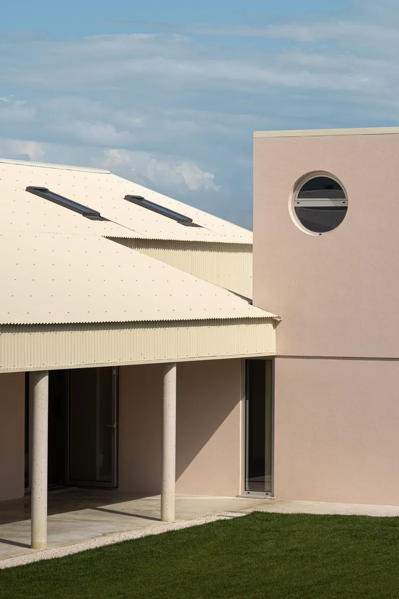 Junction between the corrugated metal roof and pink-rendered wall with circular window under afternoon light