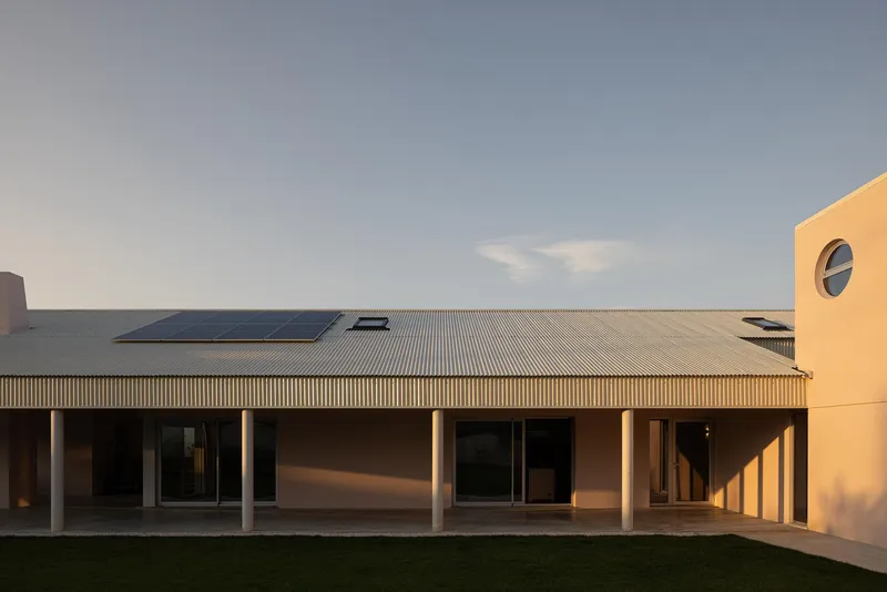 Long facade with corrugated metal roof, solar panels, and colonnade overlooking a gravel courtyard