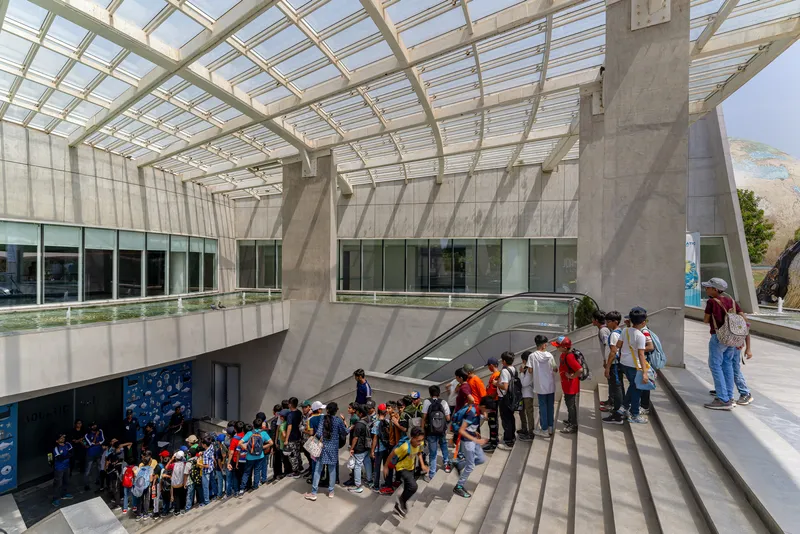 Double-height atrium with gridded glass canopy and concrete columns casting shadows over visitors queuing on an escalator