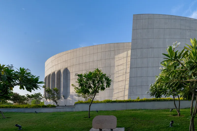 Ground-level view of the paneled stone facade with recessed arched openings behind frangipani trees and lawn