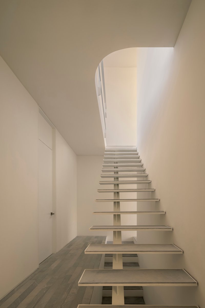 Open-riser staircase with white treads and central spine curving up toward a skylit landing