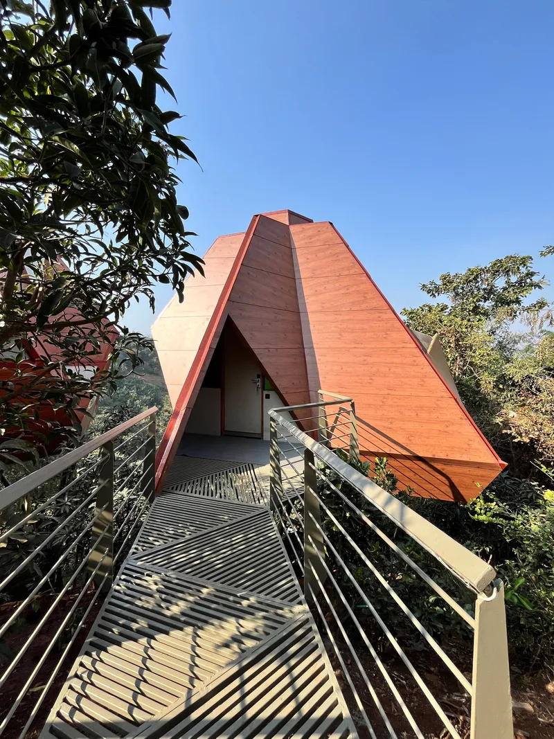Slatted metal walkway leading to a pyramidal timber-clad volume framed by foliage under clear blue sky