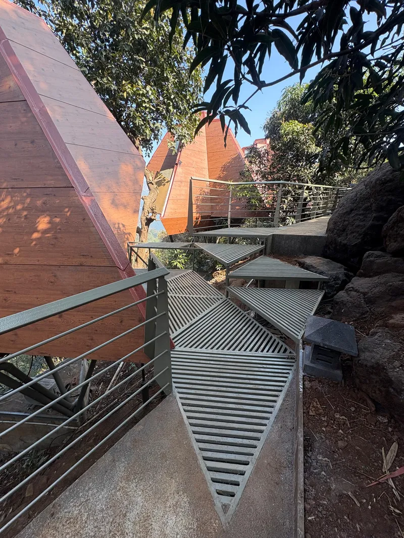 Metal walkway with horizontal railings weaving between pyramidal timber structures and boulders among trees