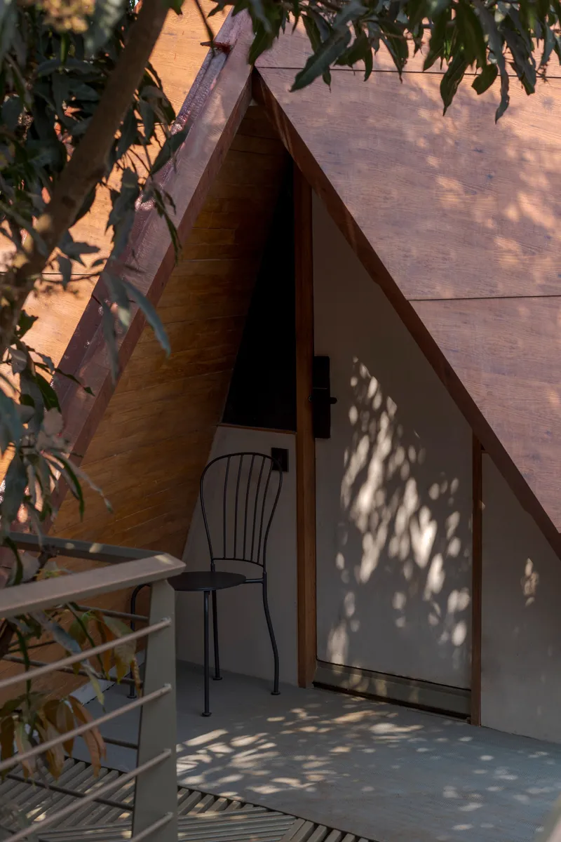 Triangular entrance framed in timber with dappled tree shadows across white wall and terrace