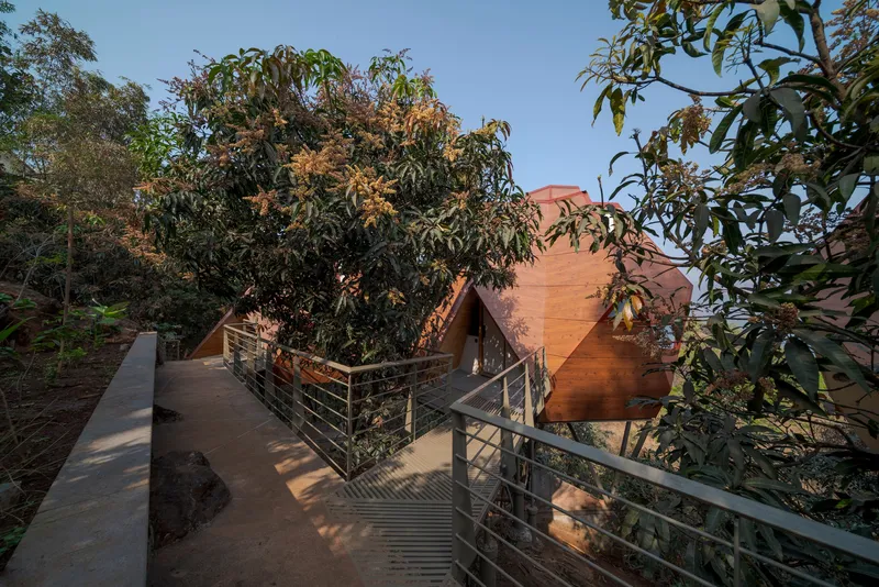 Concrete ramp with metal railings descending through vegetation toward a terracotta-clad pyramidal volume
