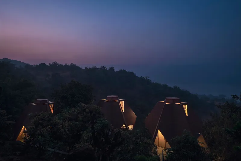 Group of triangular roof forms with glowing apertures nestled among trees at twilight