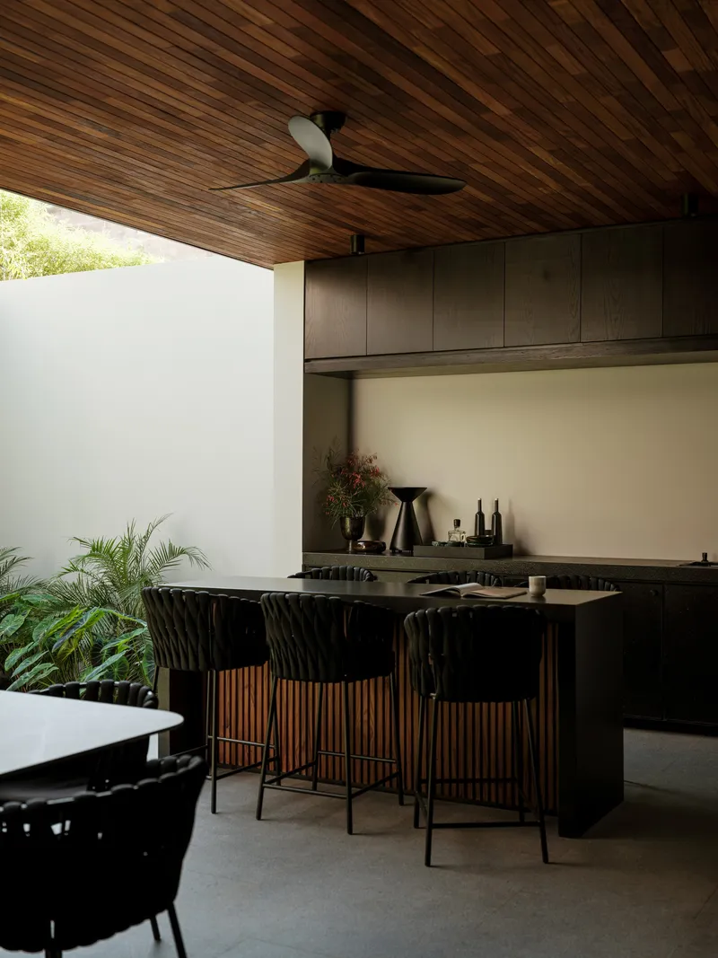 Outdoor kitchen with slatted timber ceiling, black island with bar seating, and tropical greenery beyond