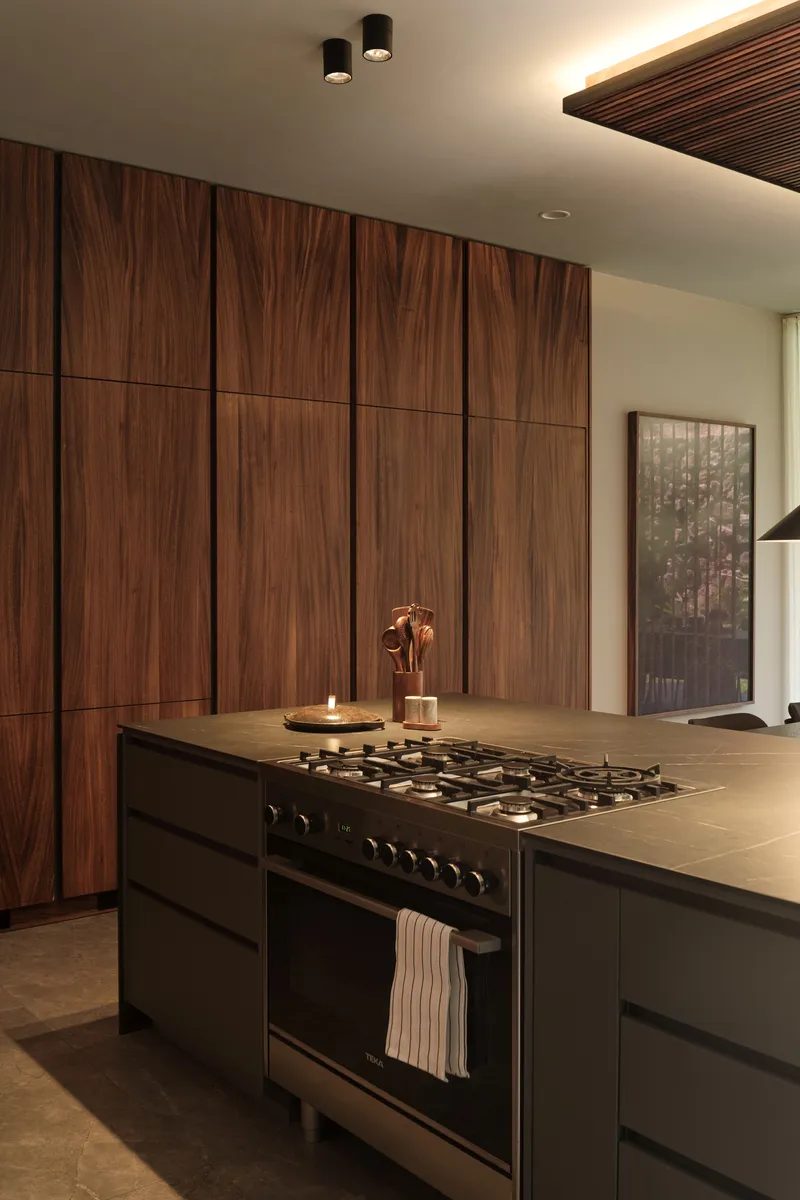 Kitchen island with black gas range against walnut veneer wall beneath recessed cove lighting
