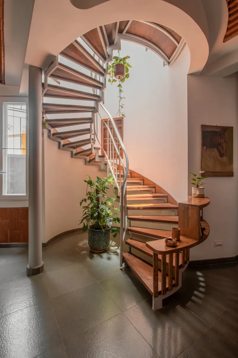 Wooden spiral staircase with open risers curving upward through a double-height white volume with potted plants