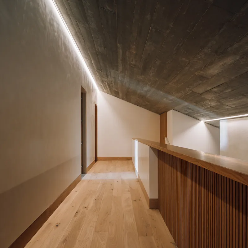 Upper corridor with timber-slatted balustrade and exposed board-formed concrete ceiling under linear skylight